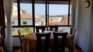Ein Essbereich mit Tisch und Stühlen, große Fenster mit Vorhängen und Blick auf Gebäude sowie einen Strand.