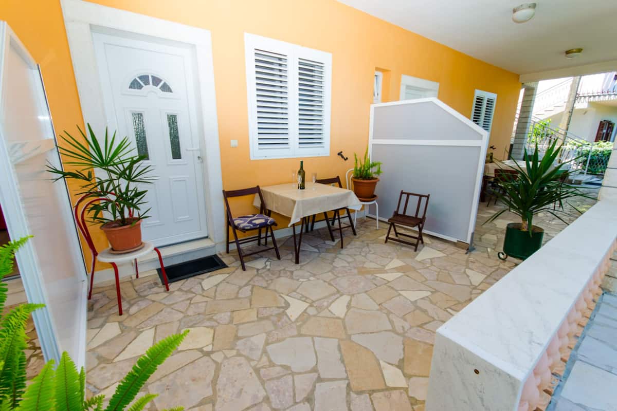Un patio con suelo de piedra, una puerta blanca y una mesa con sillas. Se ven plantas en macetas y un separador blanco.