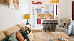 Un soggiorno moderno con un divano, un tavolino da caffè e una poltrona a fantasia. Sullo sfondo, si intravede un angolo cottura con uno sgabello da bar e un cartello con la scritta 'Cuisine'.