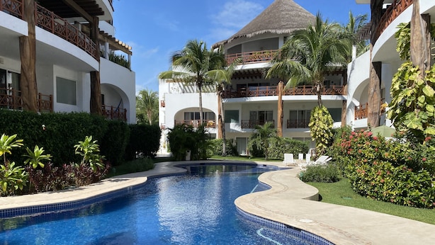 A swimming pool with a curved design, surrounded by lush greenery and palm trees. - Luxury Duplex  2 MIN to 5th , Beach & Tourist Aera (Playa del Carmen)