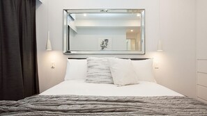 A neatly made bed with a large mirror above it, two wall-mounted lamps, and a framed picture on the wall.