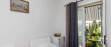 A bedroom with a bed, bedside table, and a small refrigerator. There is a balcony with a table and chairs, and a painting on the wall.