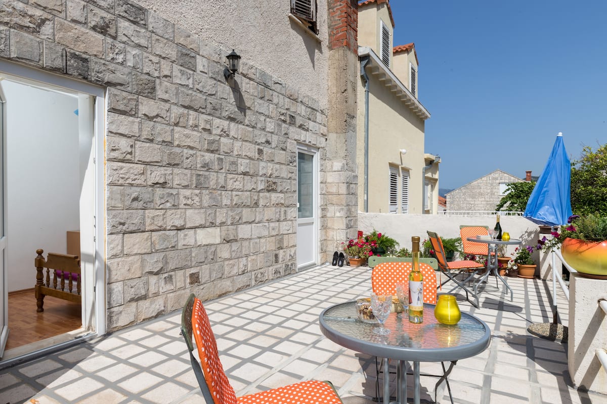 Een terras buiten met een stenen muur, een glazen tafel en oranje stoelen.