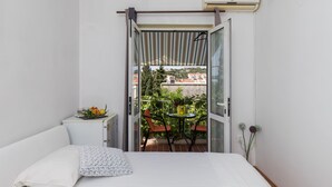 Une chambre à coucher avec un lit, une petite table sur laquelle se trouvent un vase et des fruits, et un balcon avec des chaises et des plantes.