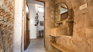 Un couloir étroit avec un mur en pierre rustique, une porte en bois et une petite étagère sur laquelle se trouvent un miroir et un robinet en laiton.