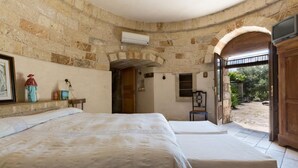 Una stanza con pareti in pietra, un letto, un tavolino con una lampada e una porta aperta che dà sull'esterno.