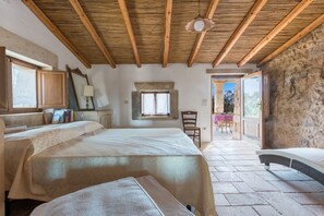 A bedroom with a large bed, wooden ceiling beams, a stone wall, and a view of the outside.