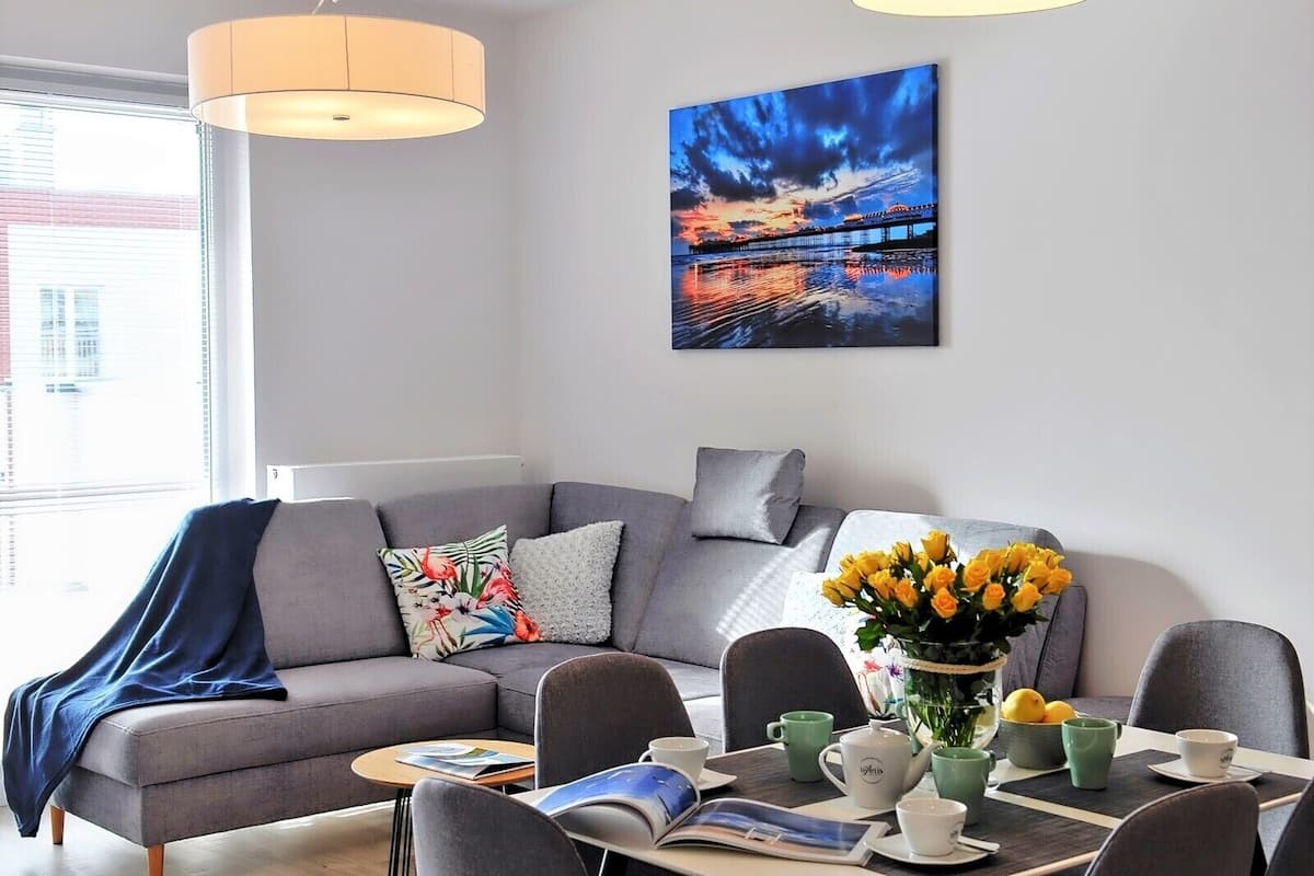 Ein modernes Wohnzimmer mit einem grauen Sofa, einem Couchtisch und einem Essbereich mit Stühlen und einer Vase mit gelben Blumen.