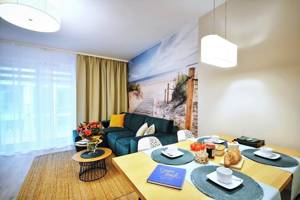Ein gemütliches Hotelzimmer mit einem Meer-Mural, einem Esstisch gedeckt mit Tellern, Tassen und einem Buch mit dem Titel "Guest Book".