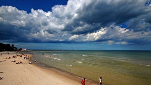 A beach with people sunbathing and swimming, a lighthouse in the background, and a cloudy sky. - BalticON Nadmorskie Tarasy (Kolobrzeg)