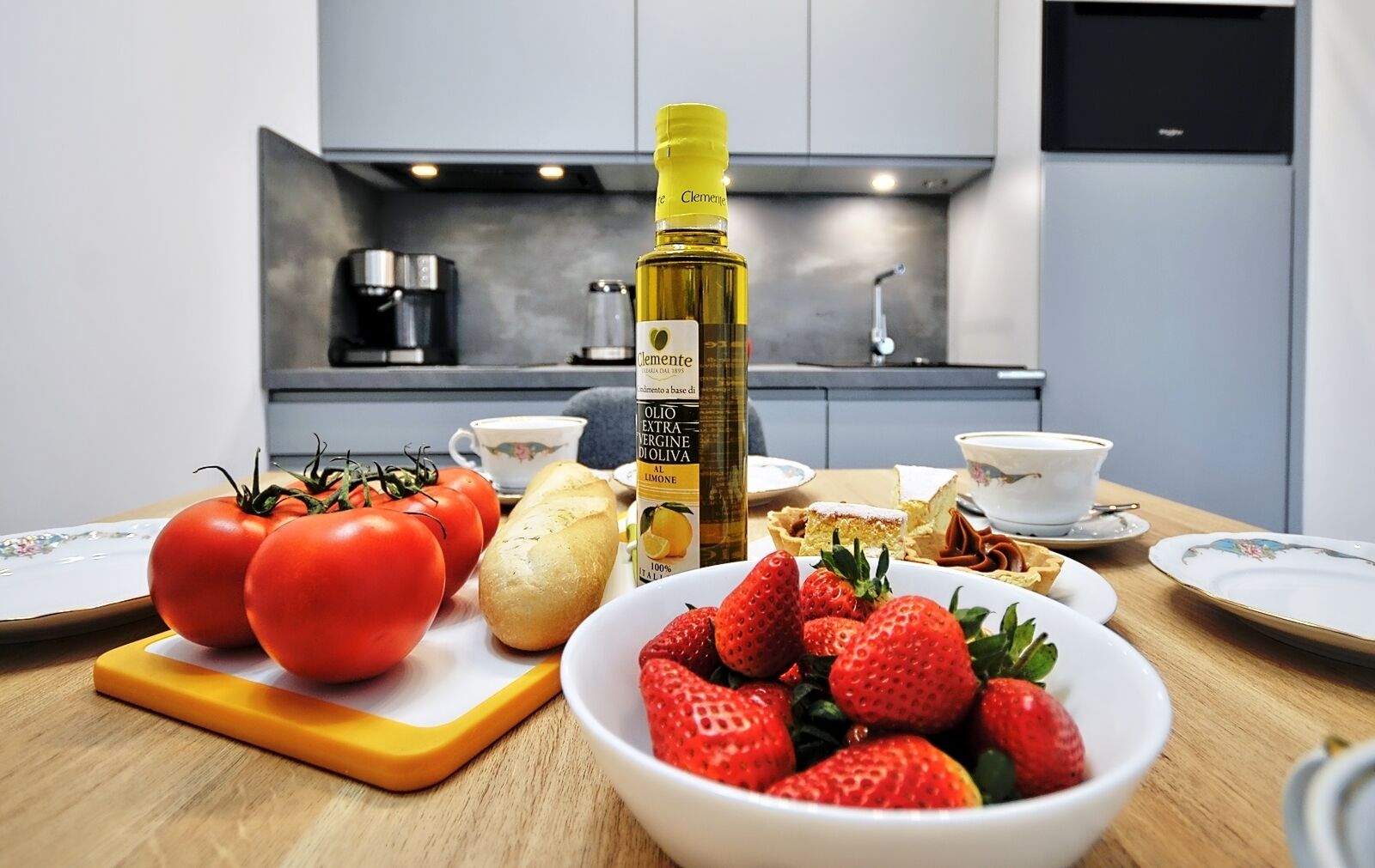 A kitchen with a table set for a meal, including tomatoes, bread, strawberries, and a bottle of olive oil.