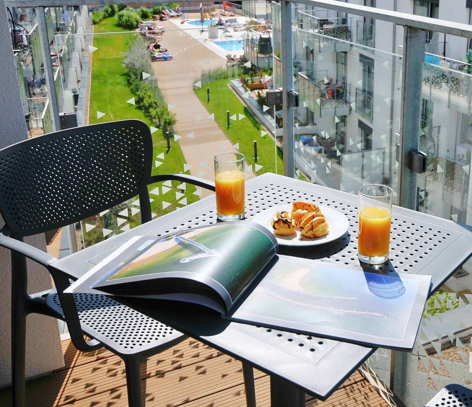 Un balcon avec une table préparée pour le petit-déjeuner, comprenant des croissants et du jus d’orange, donnant sur une piscine et un jardin.