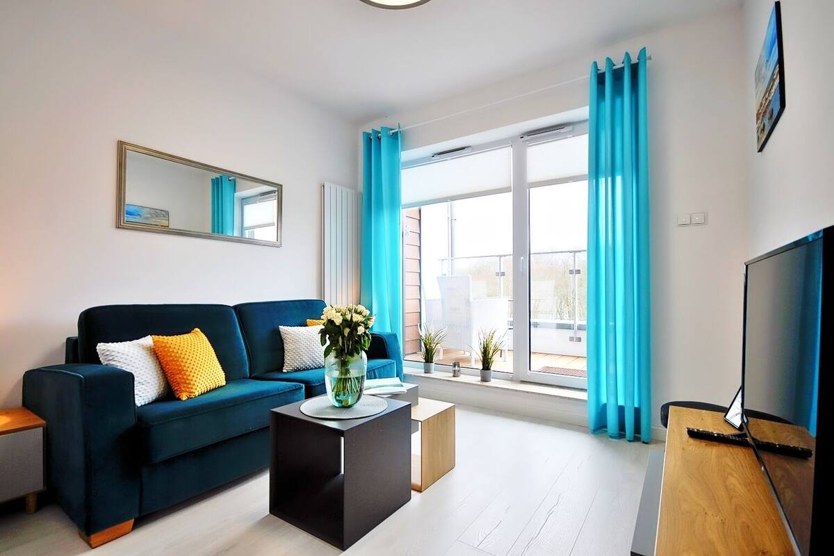 Ein modernes Wohnzimmer mit einem dunkelblauen Sofa, einem Couchtisch und einem Fernseher auf einem Holzboden. Ein großes Fenster mit blauen Vorhängen bietet einen Blick nach draußen.
