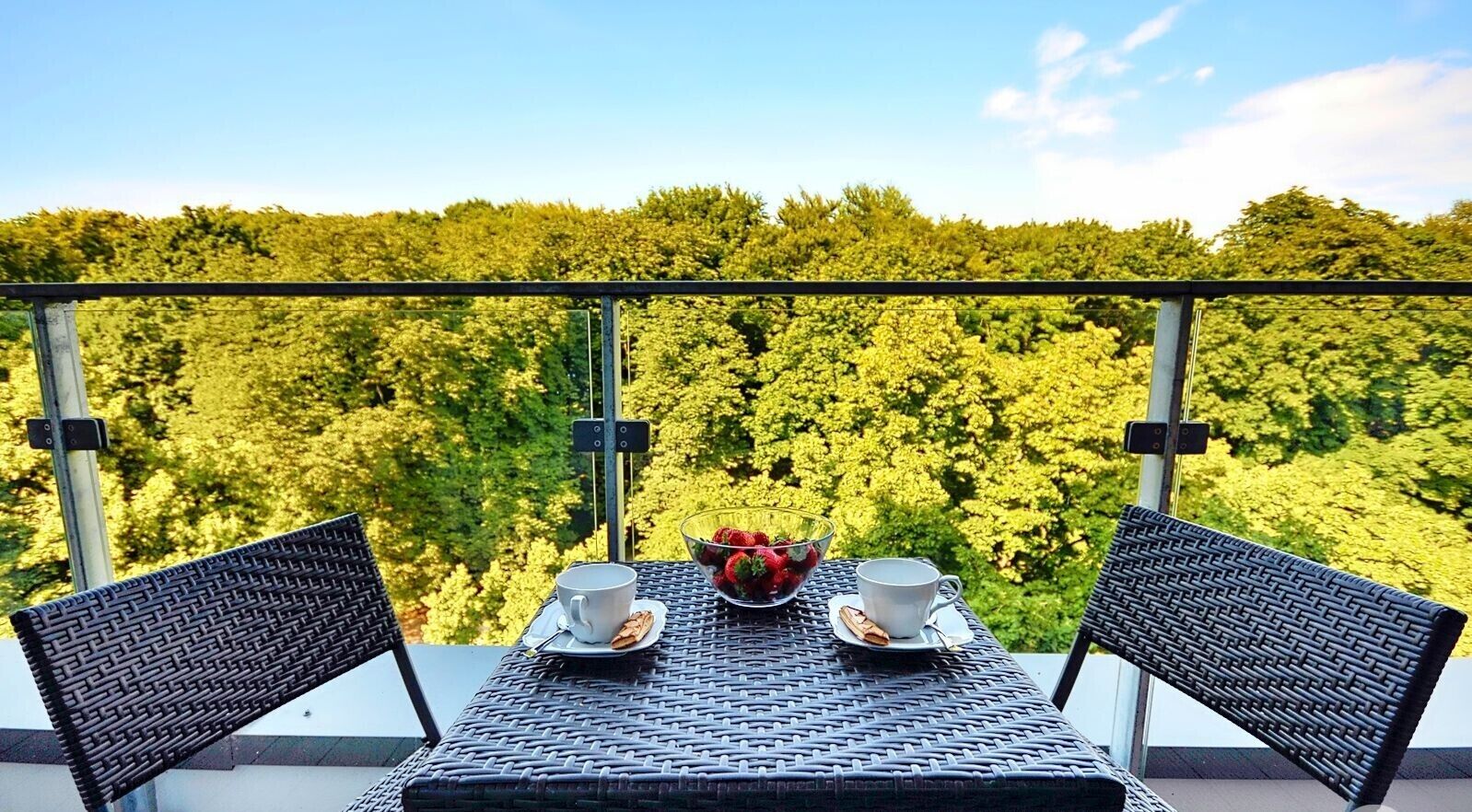 En balkon med fletstole og et bord dækket til to, med udsigt over en tæt skov.