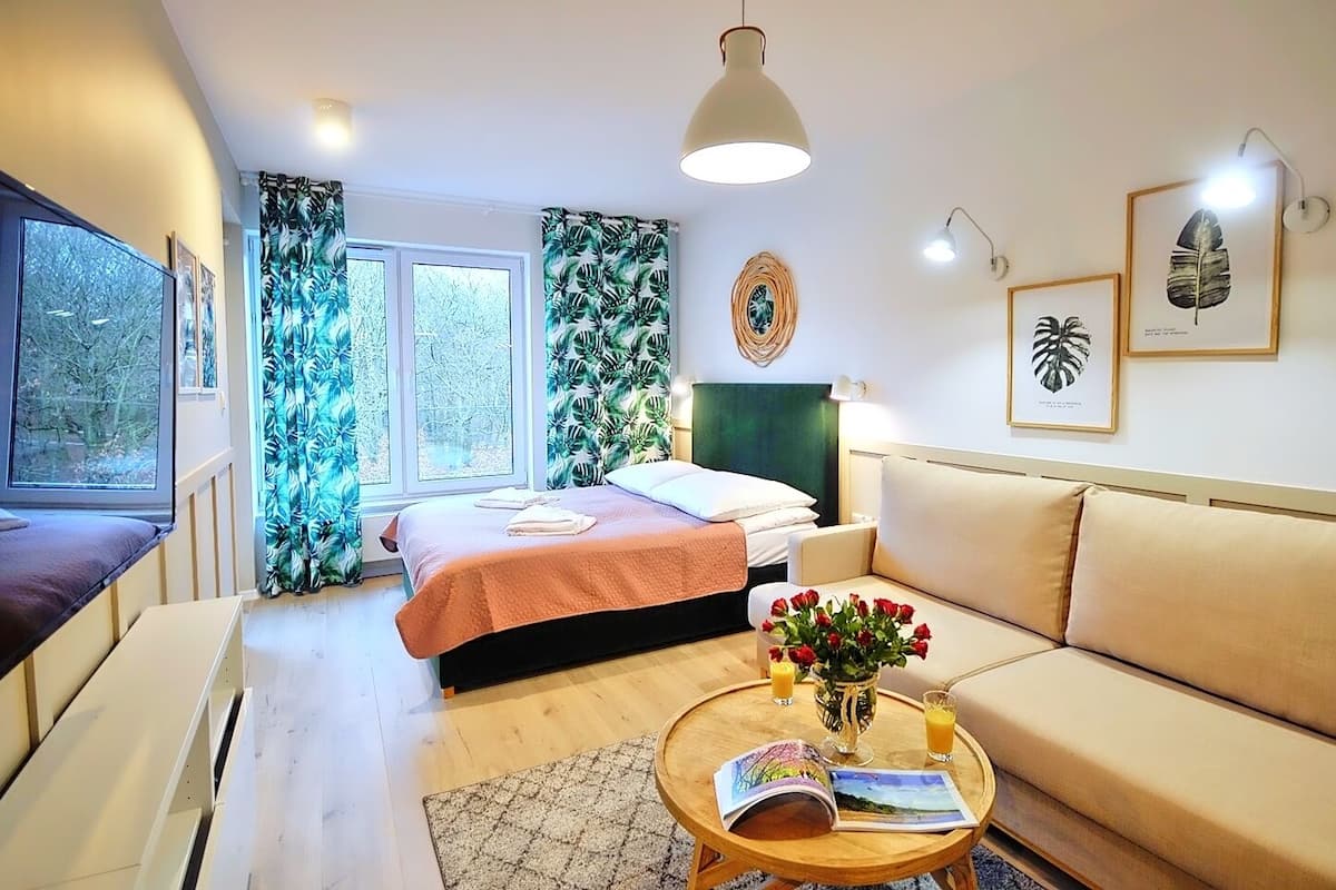 Ein gemütliches Zimmer mit einem Bett, einem Sofa, einem Couchtisch mit Buch und Blumen sowie einem Fenster mit Vorhängen.