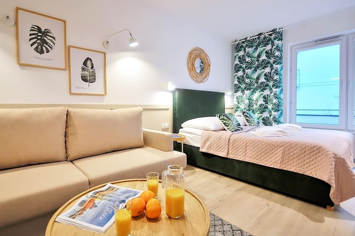 Ein modernes Hotelzimmer mit einem beigen Sofa, einem Bett mit einem grünen Kopfteil, einem runden Holztisch mit Orangen und Saft sowie Vorhängen mit tropischem Dekor.