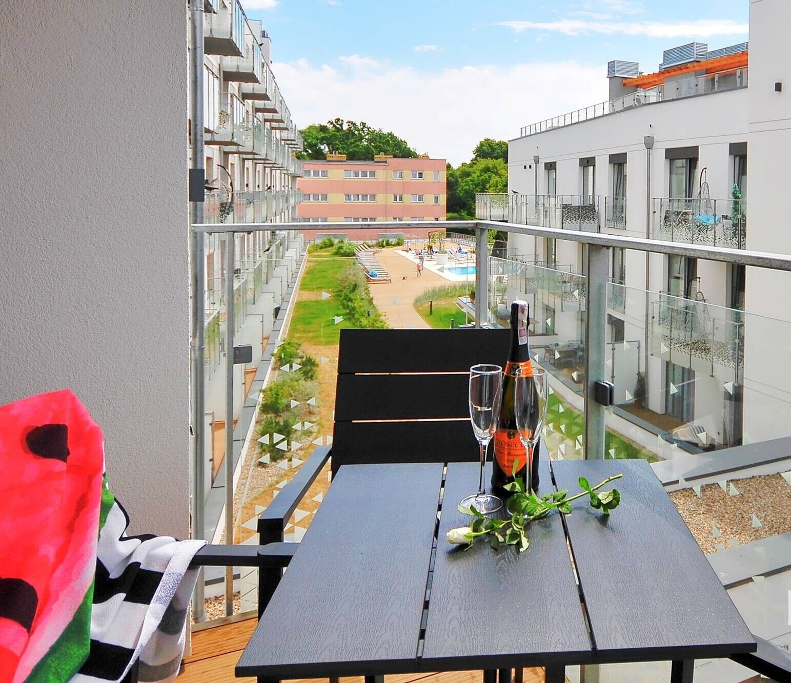 Un balcon avec une table dressée pour deux personnes, une bouteille de vin rosé et un verre. Le balcon donne sur une piscine et un jardin.