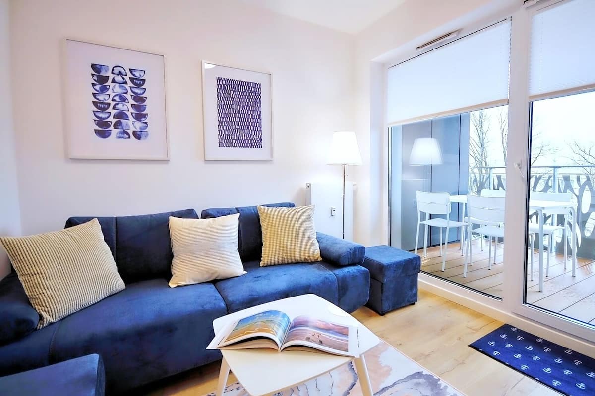 Ein modernes Wohnzimmer mit einem blauen Sofa, zwei Kissen, einer Stehlampe und einem großen Fenster mit Blick auf Gartenmöbel.