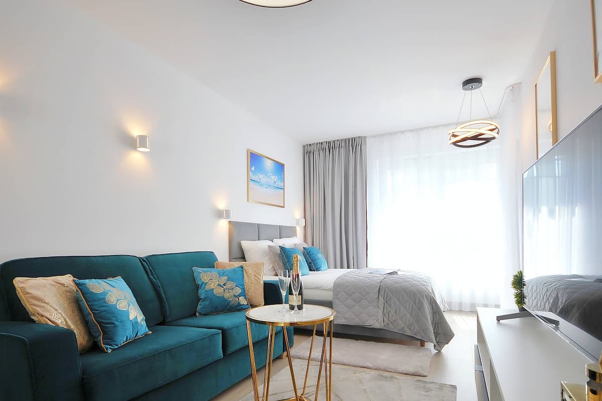 Ein modernes Schlafzimmer mit einem petrolfarbenen Sofa, einem Bett mit grauer Bettdecke, einem kleinen runden Tisch mit einer Flasche und einem großen Spiegel.
