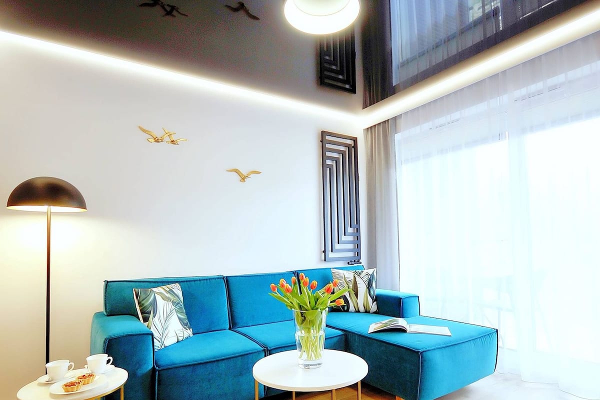 Ein modernes Wohnzimmer mit einem blauen Sofa, einem runden Couchtisch und einer Stehlampe. An der Wand befindet sich eine Dekoration und ein Fenster mit Vorhängen.