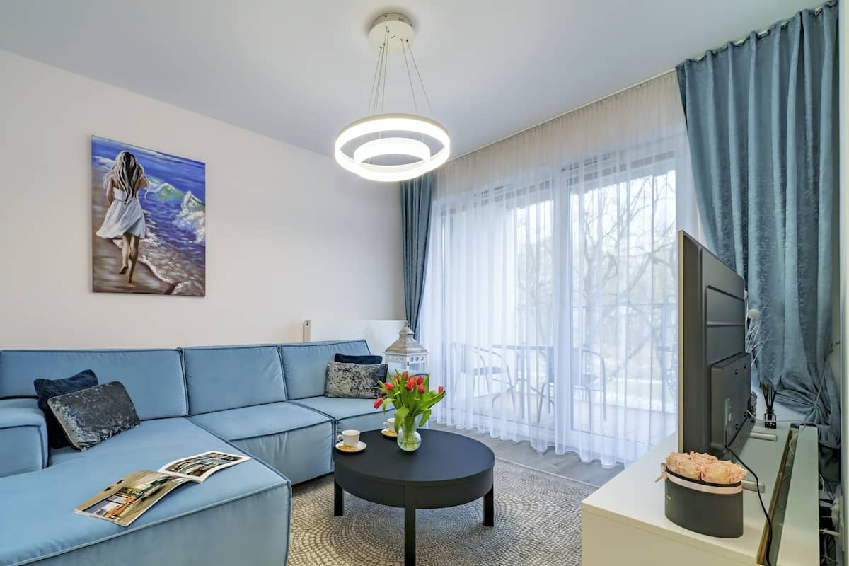 Ein modernes Wohnzimmer mit einem blauen Sofa, einem runden Couchtisch, einem Bild einer Frau am Strand und einem großen Fenster mit Vorhängen.