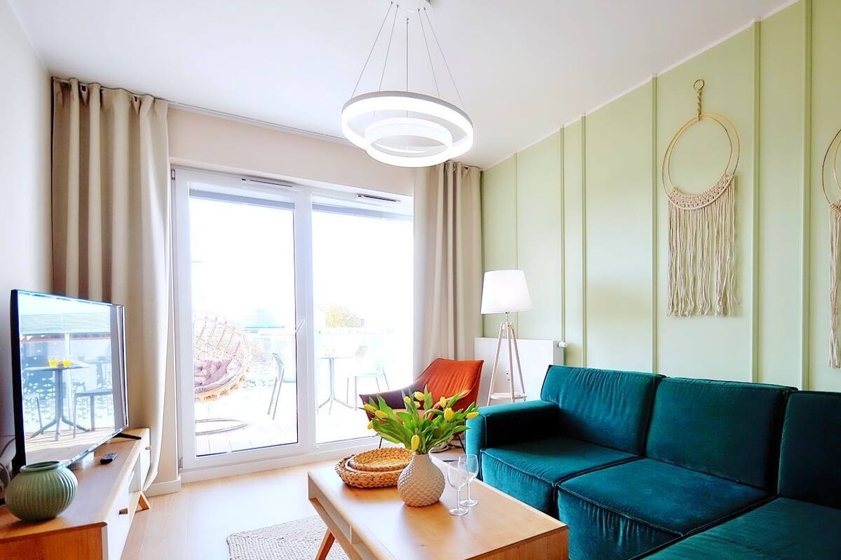 Ein Wohnzimmer mit einem salbeigrünen Sofa, einem Couchtisch mit einer Vase und Gläsern, einem Fernseher und einem großen Fenster mit Vorhängen.