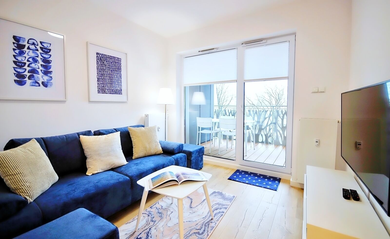 Een moderne woonkamer met een blauwe bank, een salontafel en een televisie. Twee kunstwerken in lijsten hangen aan de muur en een glazen schuifdeur leidt naar een buitenruimte.