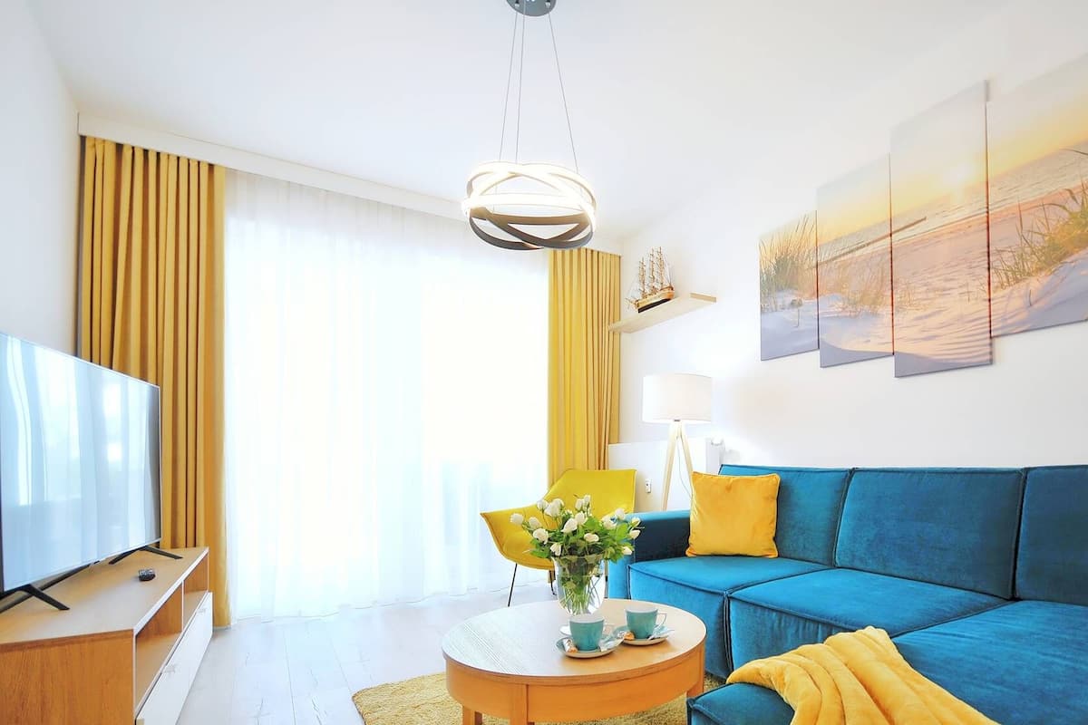 Ein modernes Wohnzimmer mit einem blauen Sofa, gelben Kissen, einem Couchtisch mit Tassen, einem Fernseher auf einem Schrank und einem großen Fenster mit Vorhängen.
