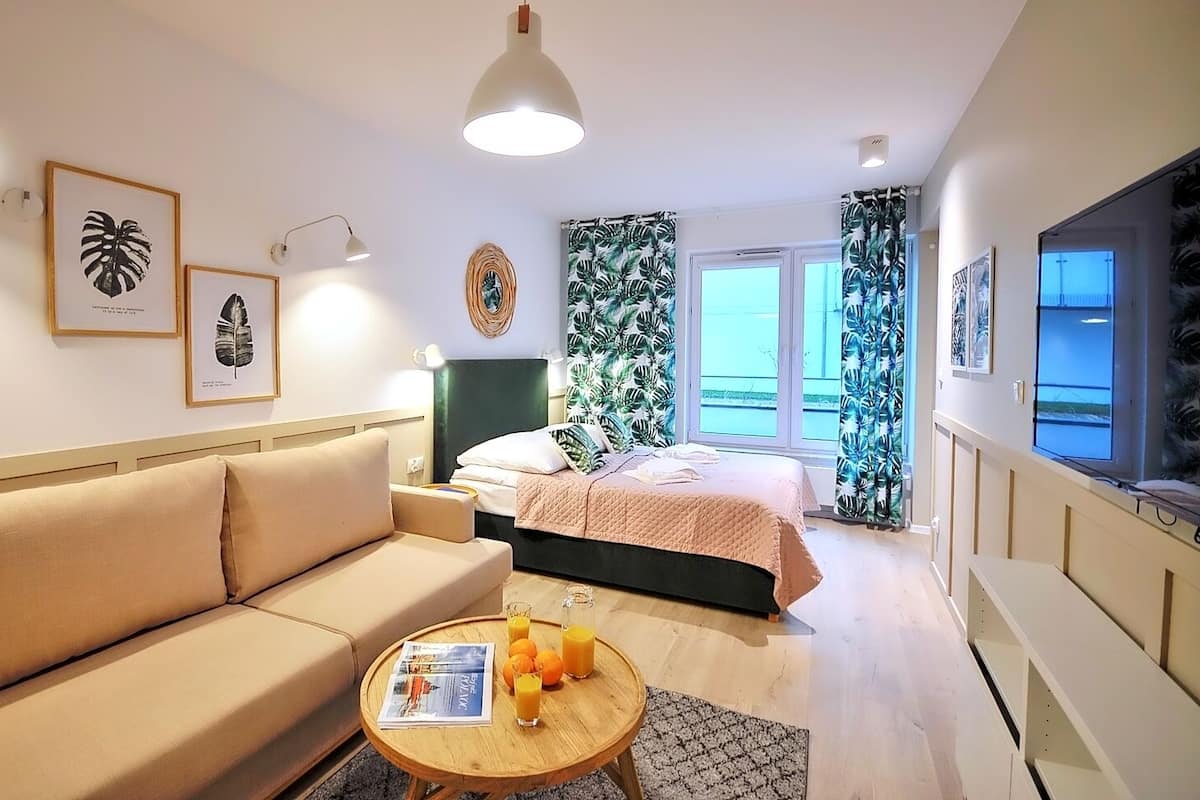 Ein modernes Hotelzimmer mit einer Couch, einem Bett, einem kleinen Tisch mit Zeitschrift und Orangensaft sowie einem Fernseher auf der rechten Seite.