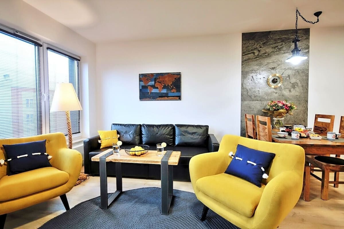 Ein Wohnzimmer mit einer Couch, zwei gelben Sessel, einem Couchtisch und einem Essbereich mit Tisch und Stühlen.