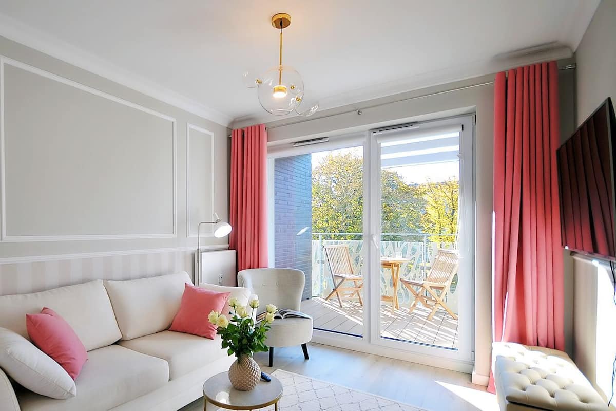 Ein modernes Wohnzimmer mit einer Couch, einem Couchtisch und einem Sessel. Es gibt einen Balkon mit Blick auf Bäume und ein blaues Gebäude.