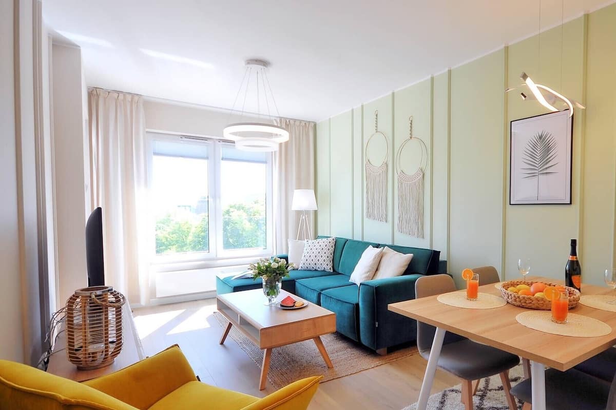 Ein modernes Wohnzimmer mit einem türkisgrünen Sofa, einem Holzespeisetisch mit Platzsets, einem gelben Sessel und einem großen Fenster mit Vorhängen.