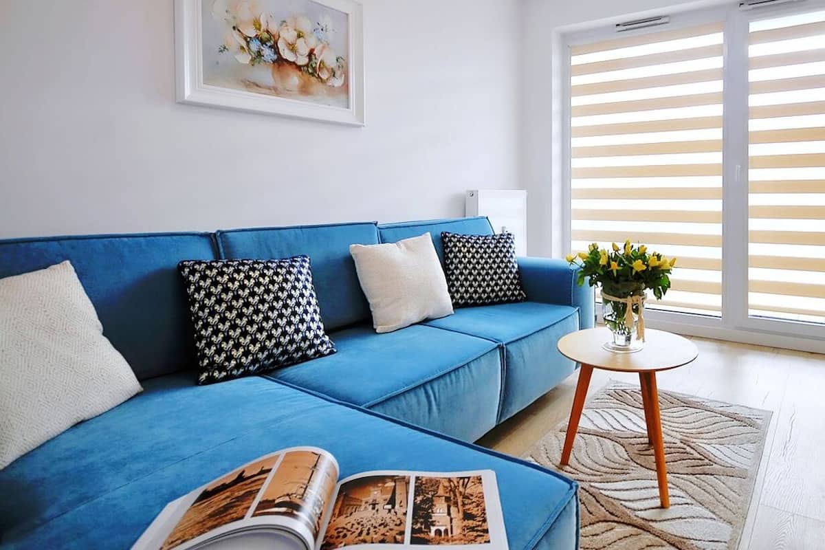 Ein modernes Wohnzimmer mit einem blauen Sofa, gemusterten Kissen, einem gerahmten Kunstwerk an der Wand und einem Fenster mit Jalousien.