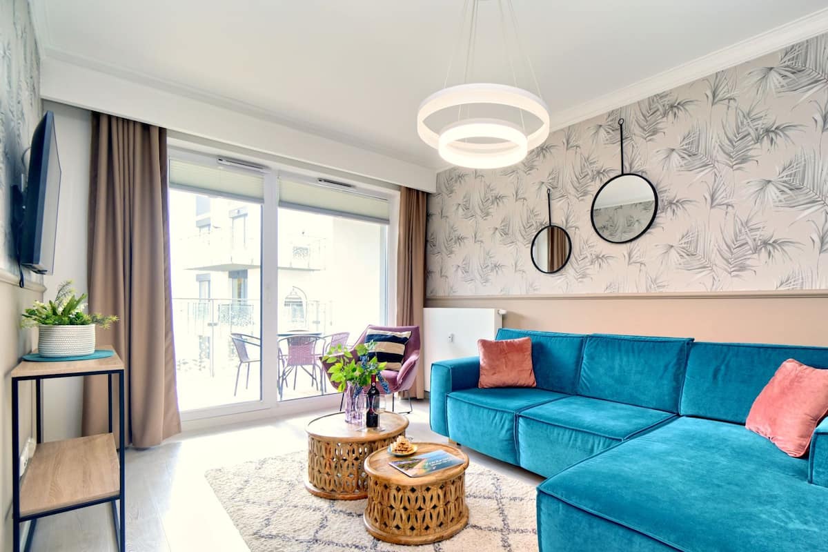 Ein modernes Wohnzimmer mit einem blauen Sofa, zwei runden Holztischen, einem großen Fenster mit Blick auf Balkone und einer Wand mit einem Blattmuster.