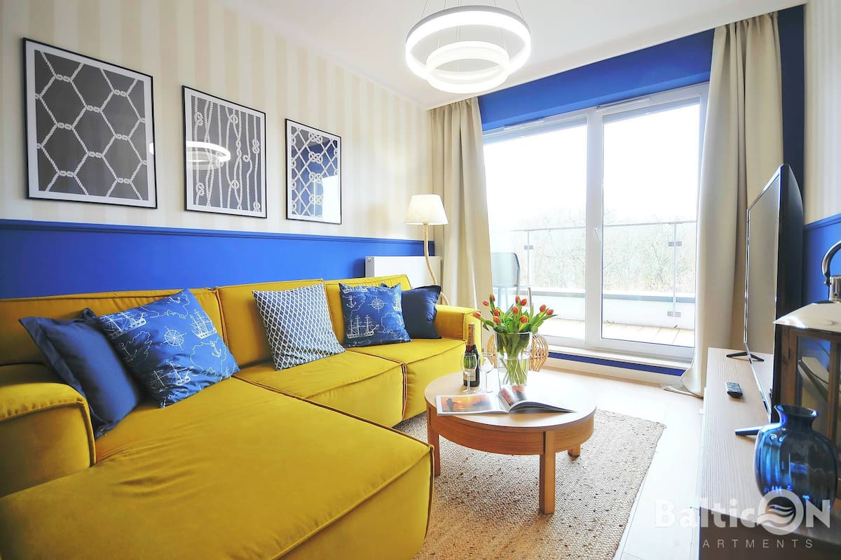 Ein helles Wohnzimmer mit einem gelben Sofa, einem runden Couchtisch und einem großen Fenster mit Vorhängen.
