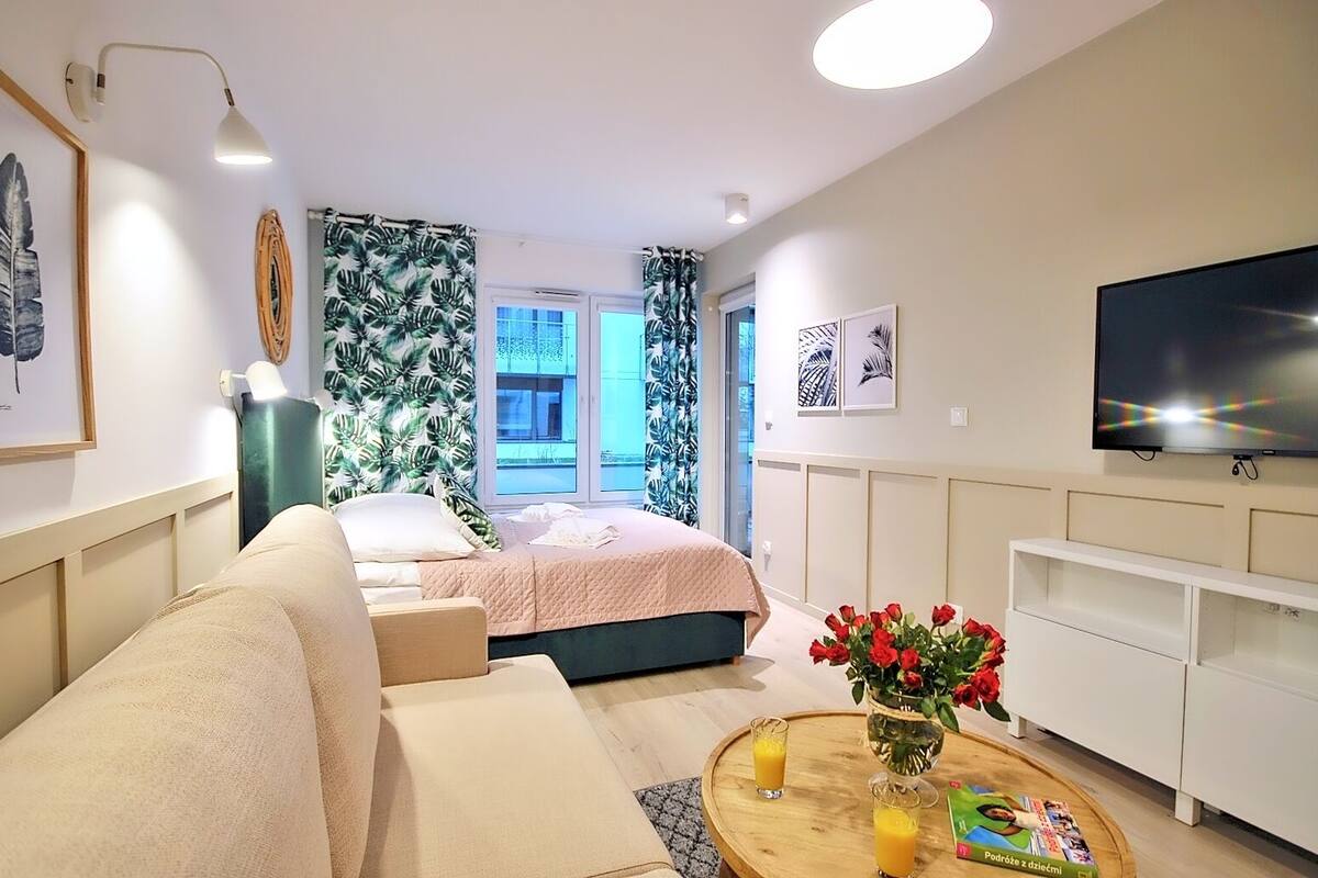 Ein moderner, gut beleuchteter Raum mit einem Bett, einer Couch, einem Fernseher und einem Couchtisch mit einem Buch und Getränken.