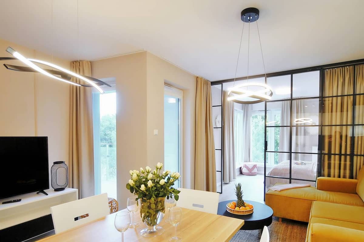 Ein modernes Wohnzimmer mit einer Couch, einem Esstisch mit Stühlen, einem Fernseher und einem großen Fenster mit Vorhängen.