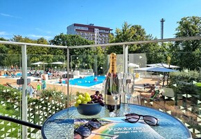 En balkong med ett glasbord, på vilket står en flaska Moët & Chandon-champagne, en skål med druvor och ett par solglasögon. I bakgrunden syns ett poolområde med folk och parasoller.