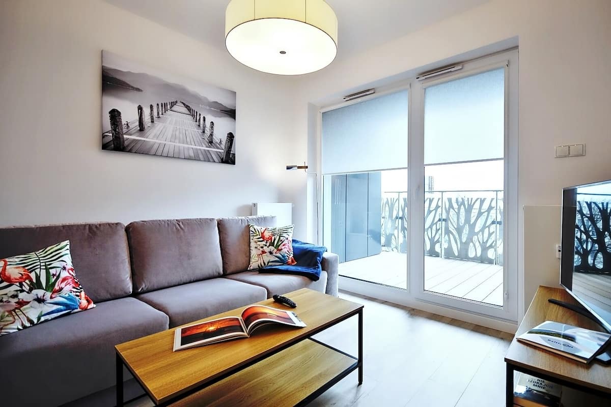 Ein modernes Wohnzimmer mit Sofa, Couchtisch und Fernseher. Ein großes Fenster bietet Blick auf einen Balkon.