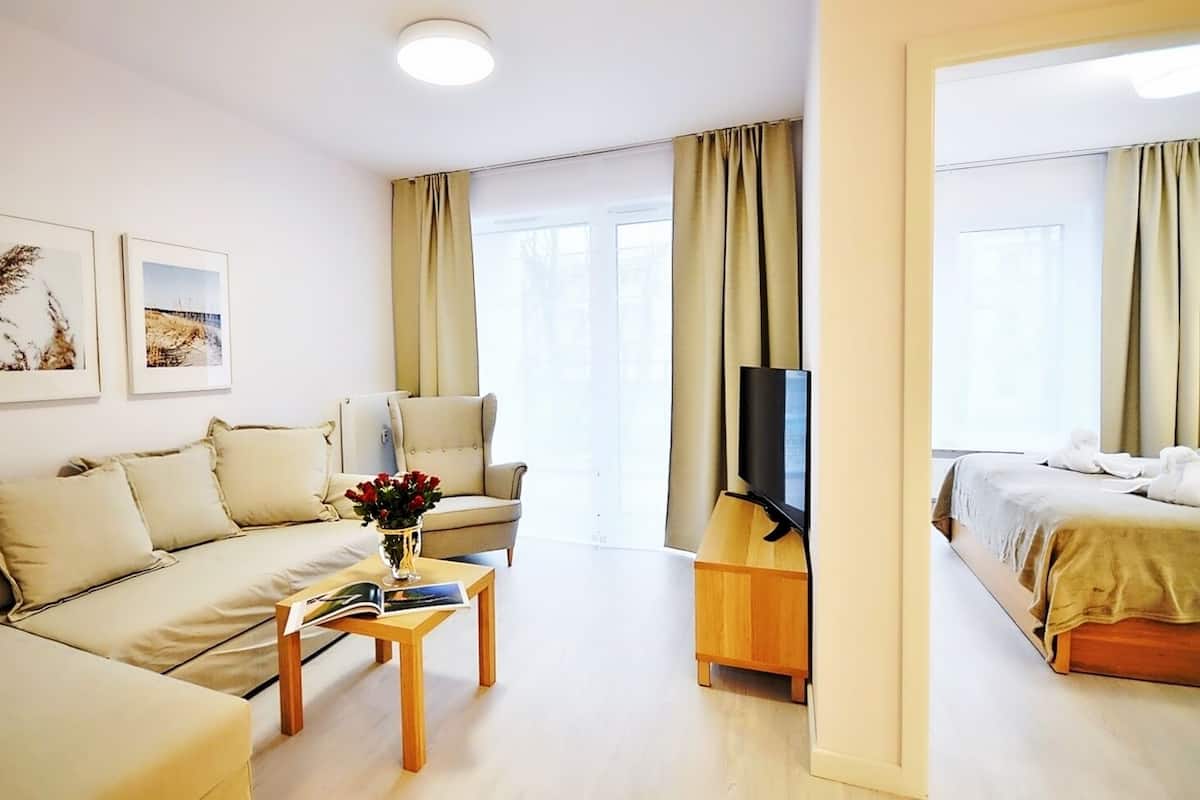 Ein modernes Wohnzimmer mit Sofa, Sessel, Couchtisch und Fernseher. Zwei Fenster mit Vorhängen sind zu sehen, und im angrenzenden Raum ist ein Bett erkennbar.
