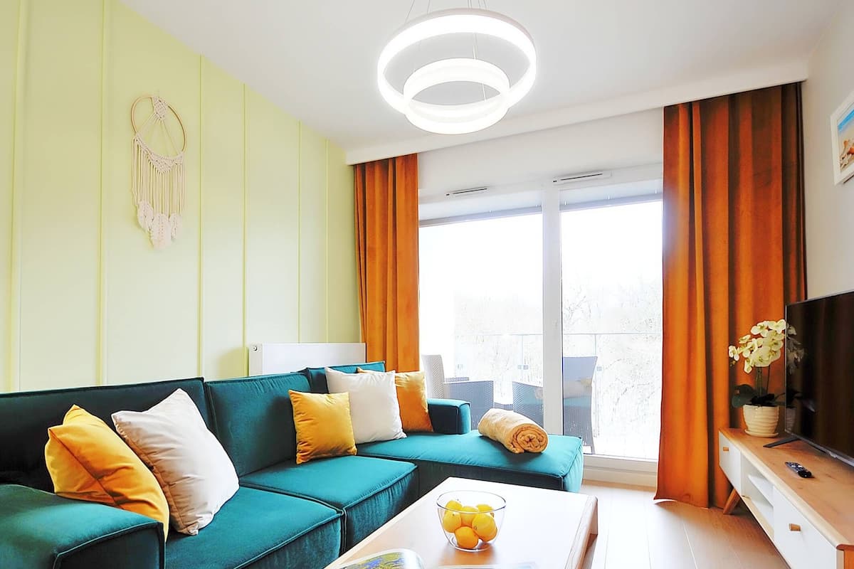 Ein modernes Wohnzimmer mit einem türkisfarbenen Sofa, orangefarbenen und weißen Kissen, einem Glastisch mit Obst, einem Flachbildfernseher und einem großen Fenster mit Vorhängen.
