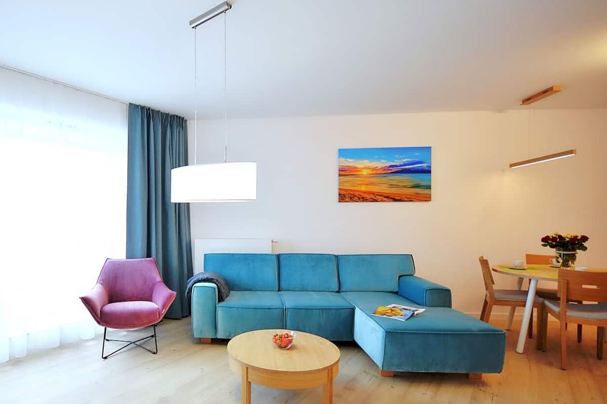 Ein modernes Wohnzimmer mit einem blauen Sofa, einem rosa Sessel, einem runden Holztisch und einem Essbereich mit kleinem Tisch und Stühlen.