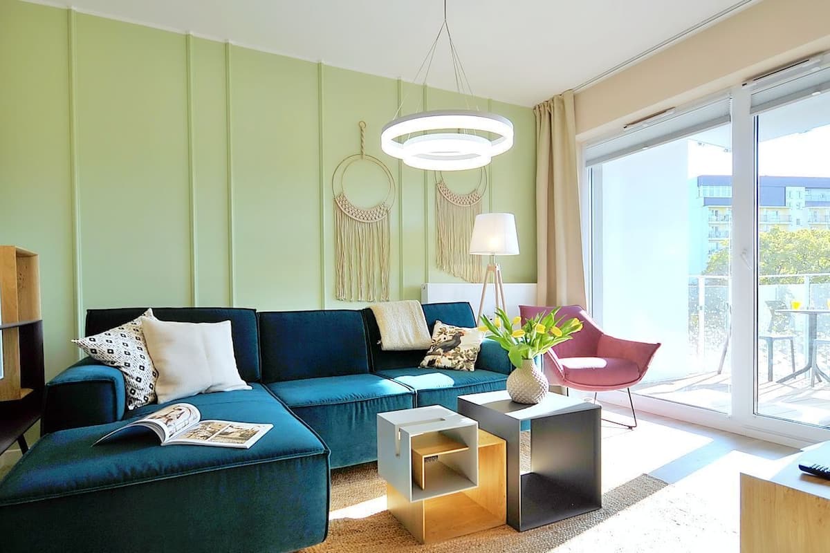 Ein modernes Wohnzimmer mit einem türkisfarbenen Sofa, einem kleinen Beistelltisch, einem roten Sessel und einem großen Fenster mit Ausblick nach draußen.