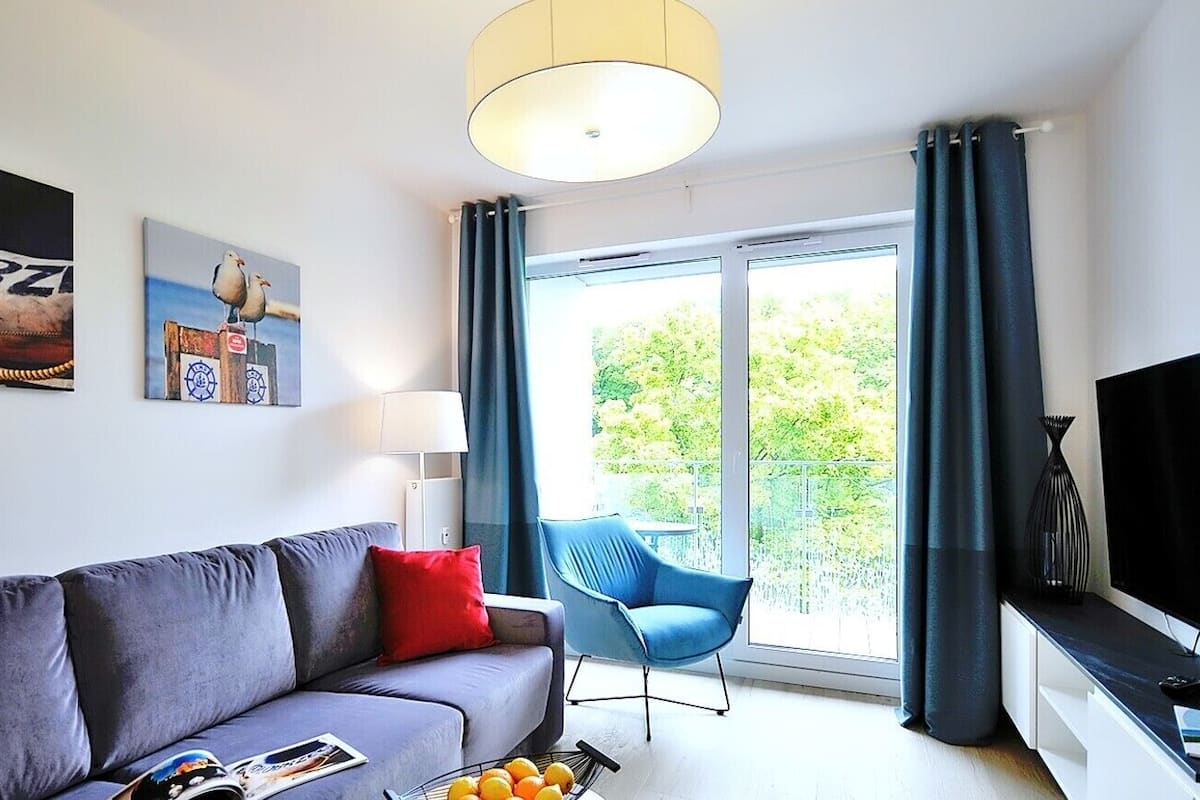 Ein modernes Wohnzimmer mit einem grauen Sofa, roten Kissen, einem blauen Sessel, einem Fernseher und einem großen Fenster mit Vorhängen.