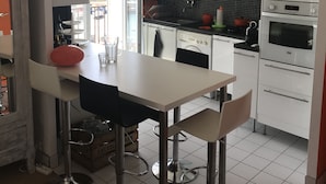 A modern kitchen with white cabinets, a stainless steel sink, and a built-in oven. There is a dining area with a white table and black chairs. - Very bright F2 with terrace and panoramic view of the Port <br> (Sète)