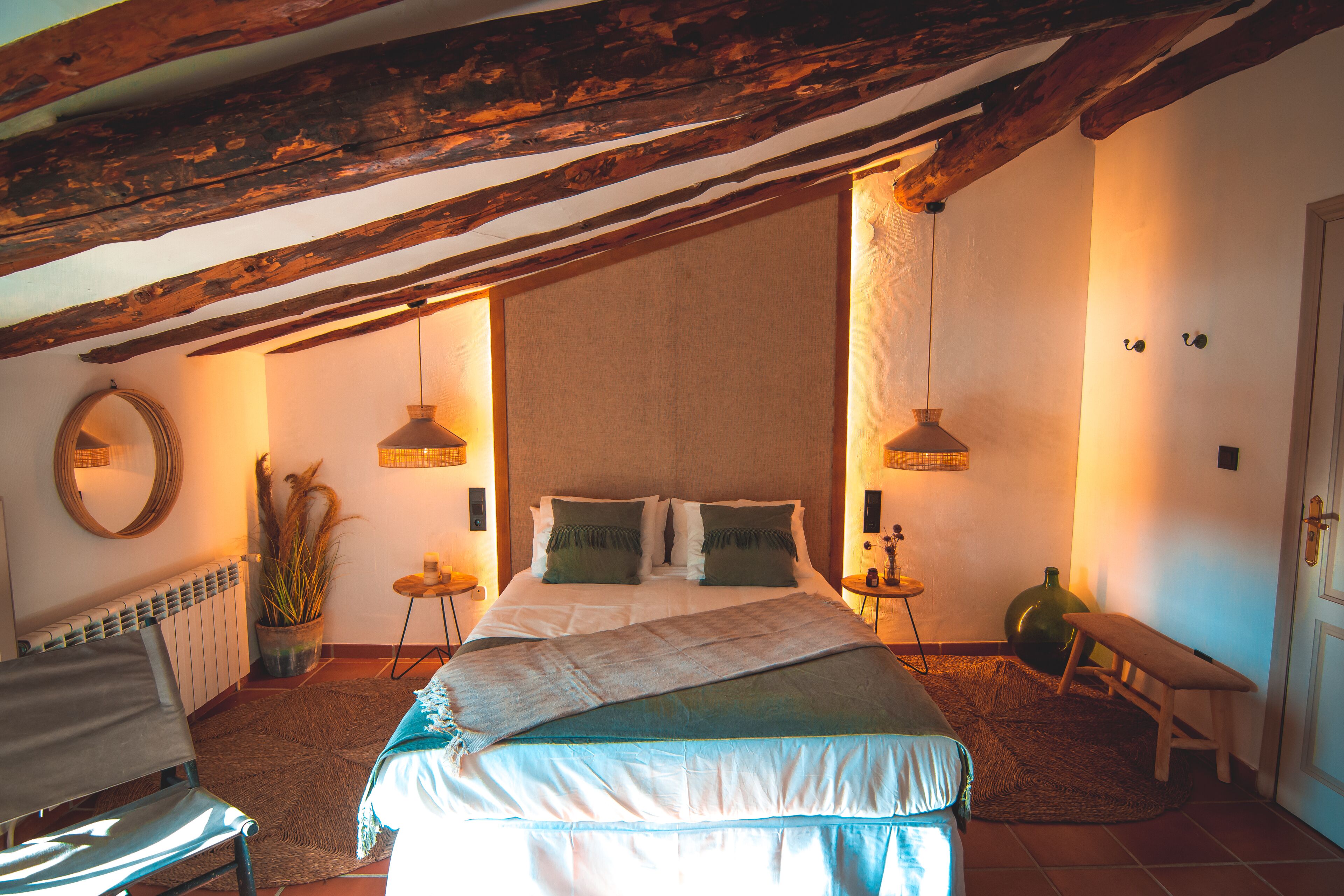 A bedroom with a large bed, wooden beams, a round mirror, two bedside tables with lamps, a bench, and a radiator.