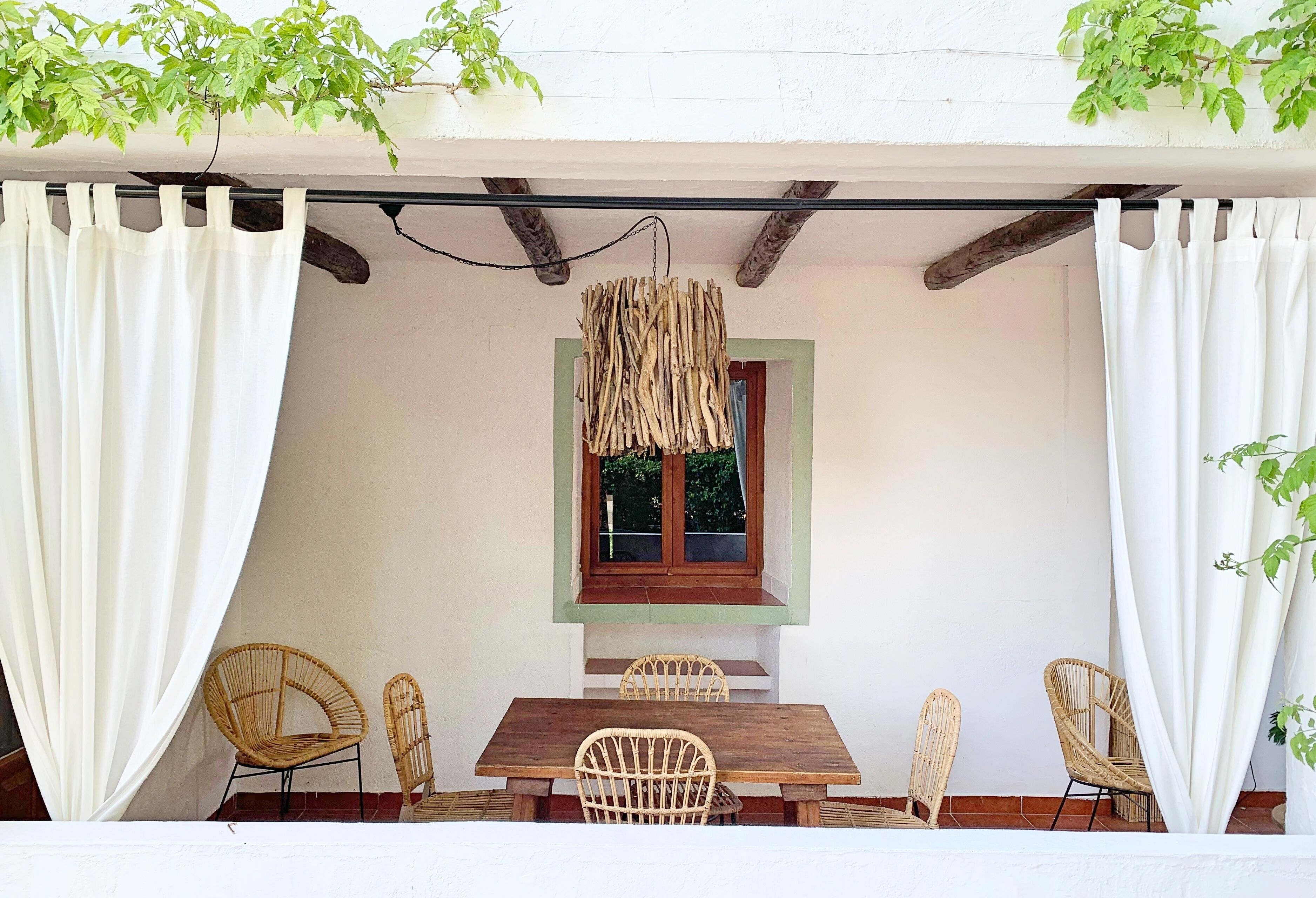 A covered patio with a wooden table and wicker chairs, a hanging light fixture, and a window with a green frame.