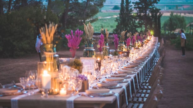 Una larga mesa para comer al aire libre, decorada con velas y cristalería, rodeada de follaje y árboles.