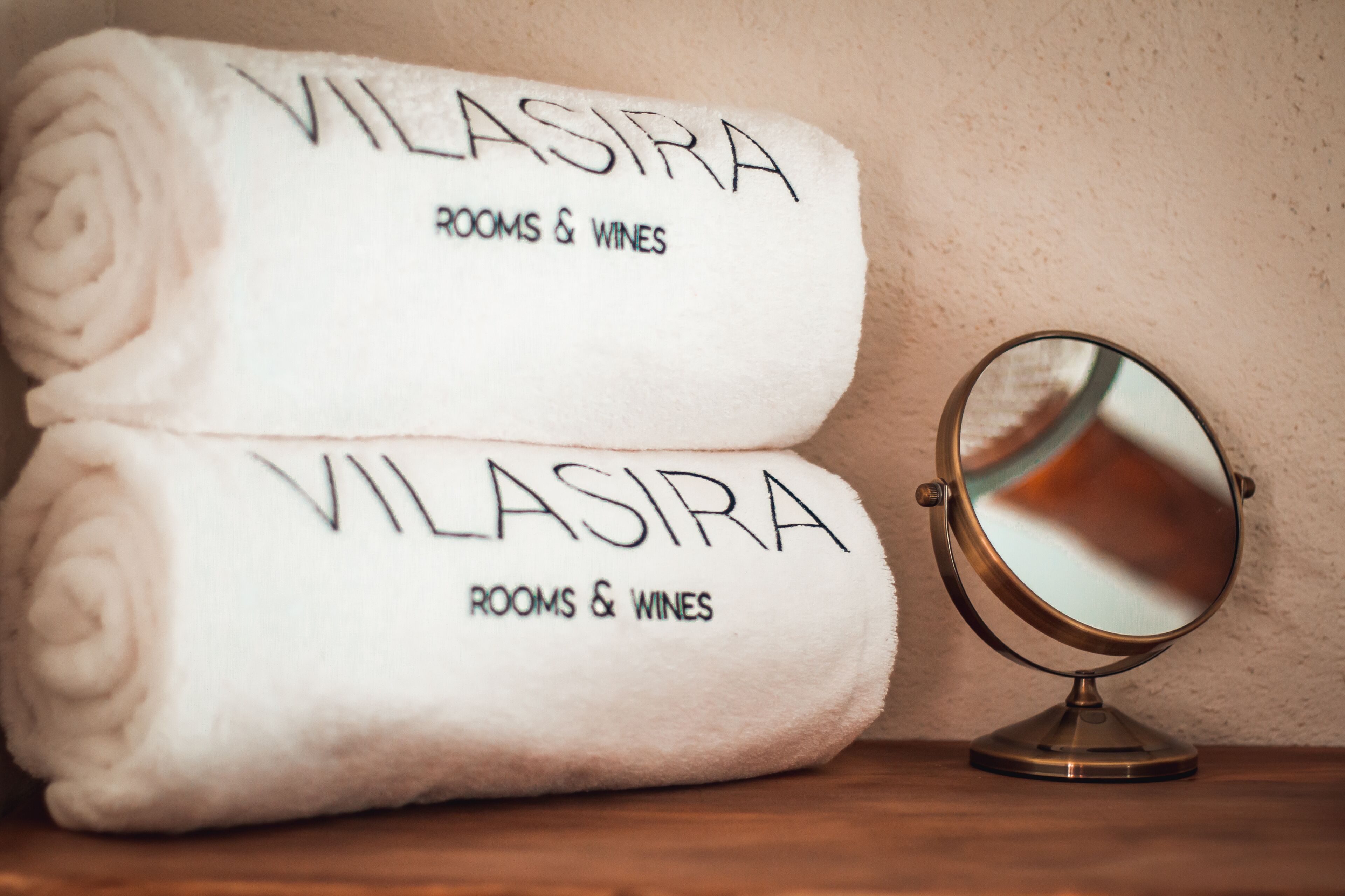 Two white towels with 'VILASIRA ROOMS & WINES' printed on them, placed on a wooden surface next to a round mirror.