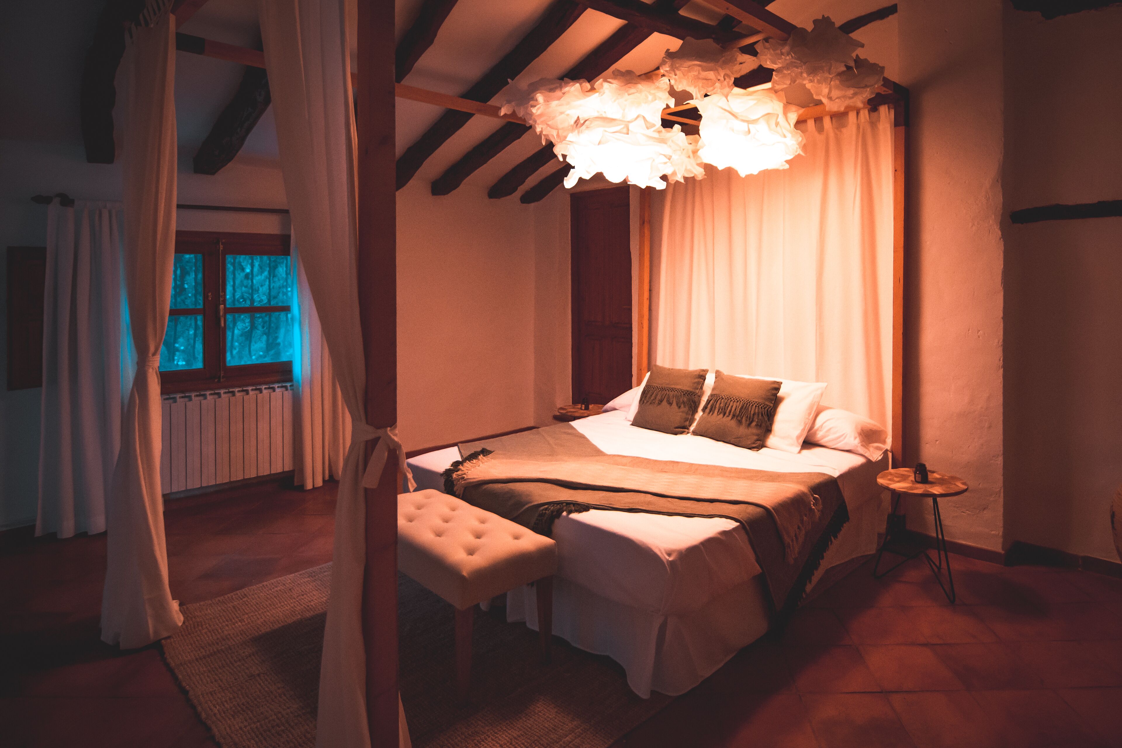 Un dormitorio con cama a dosel, un banco, una mesita y vistas a una ventana con cortinas.